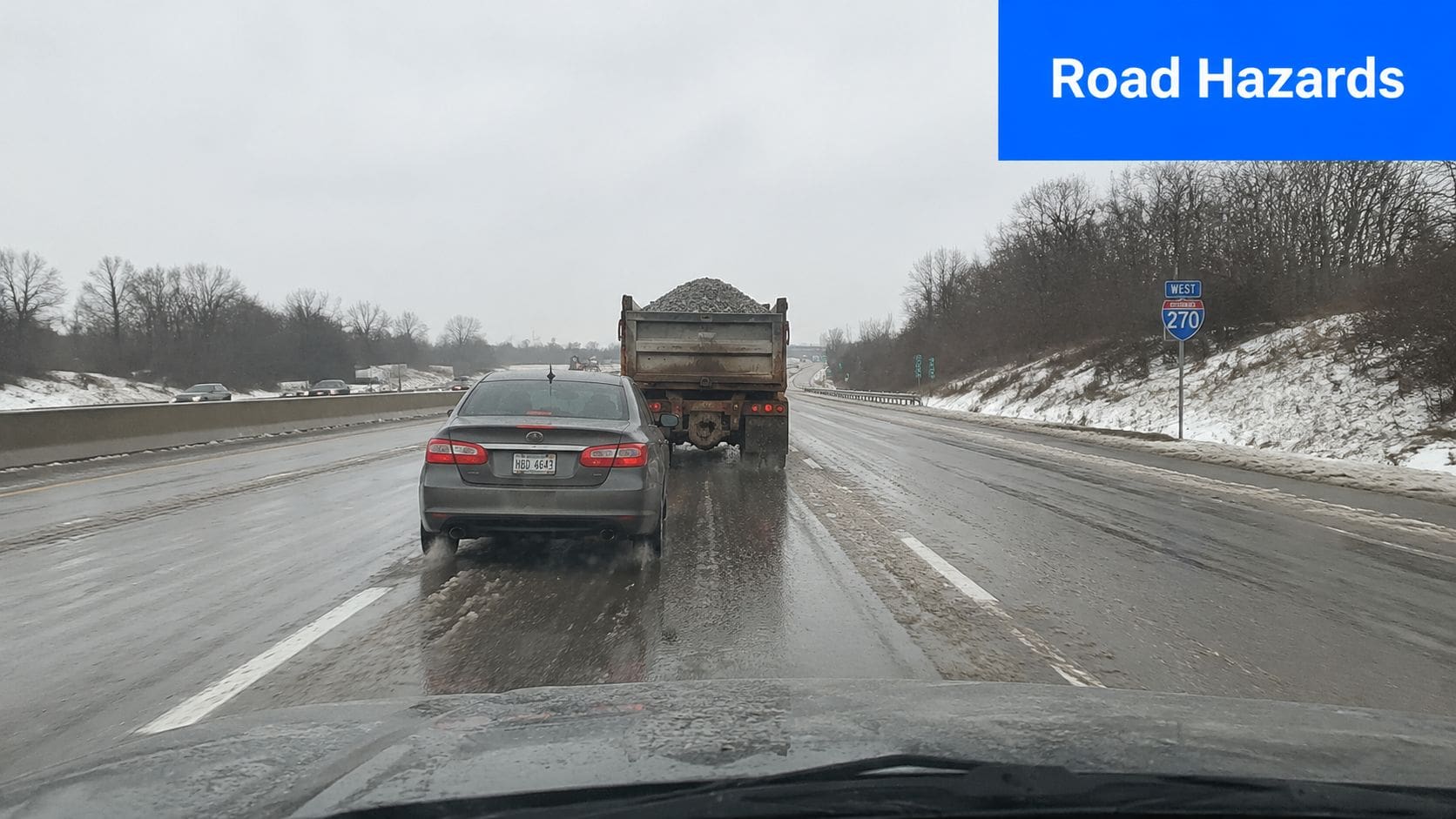 A car driving on a snowy highway behind a dump truck filled with loose rocks.
