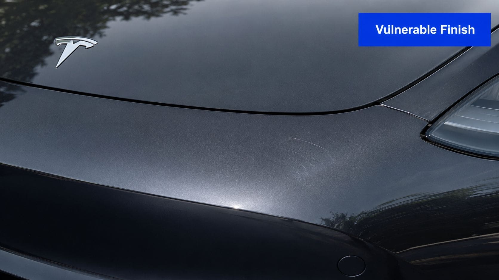 A close-up view of a dark gray Tesla hood showing visible surface scratches and swirl marks on the paint.